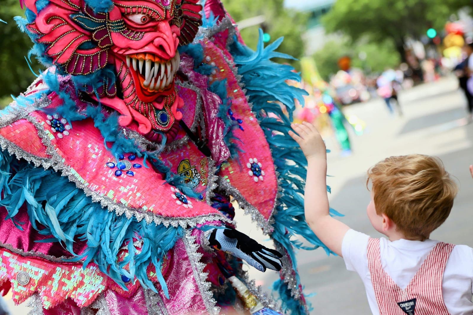 A pink and teal feathered carnival devil gives a high-five to a young boy along the parade route
