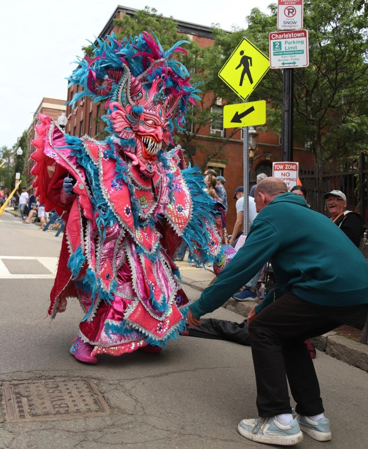 A pink and teal feathered Diablo Cojuelo performing in the street as a spectator looks on